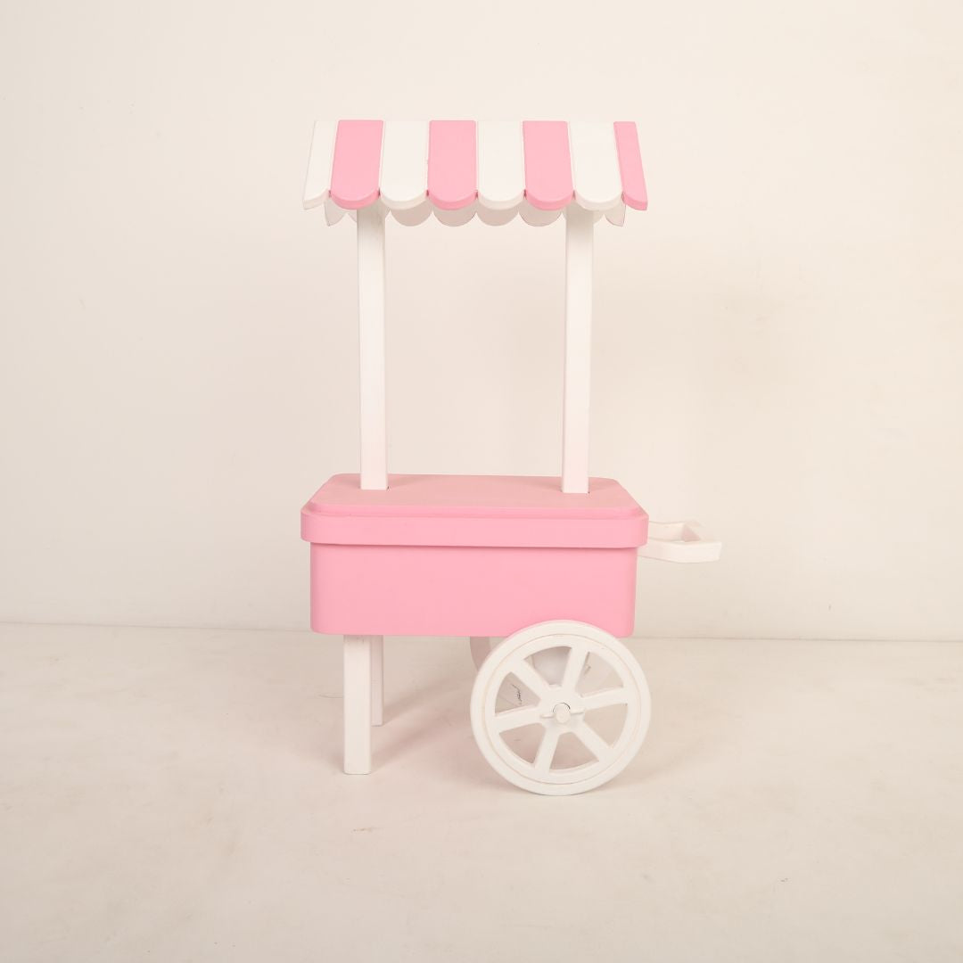 A charming candy cart baby prop designed for baby photoshoots. The cart features a pastel-colored canopy, a wooden frame with intricate details, and small decorative wheels. It is adorned with candy-themed accents, creating a whimsical and playful backdrop perfect for capturing adorable baby moments.